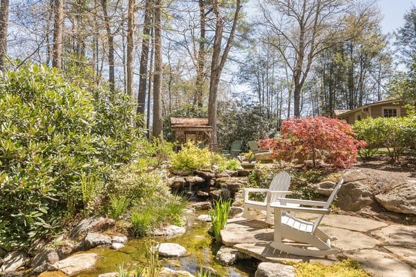 Lush garden featuring a tranquil stream, stone pathways, and seating area surrounded by mature trees.