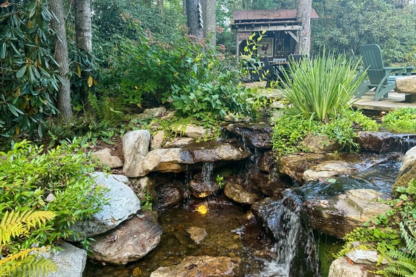 This tranquil backyard features a cascading stone waterfall, lush greenery, and a cozy retreat for relaxation.