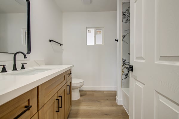 This renovated bathroom features a quartz vanity, custom cabinetry, matte black fixtures, and a striking marble-look tub and shower surround for everyday luxury.