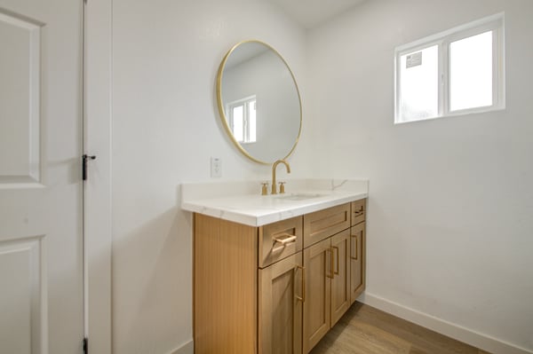 This remodeled bathroom highlights a quartz countertop, custom wood vanity, designer gold faucet, round mirror, and contemporary finishes for a polished modern look.
