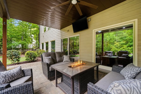 A covered outdoor living area designed for year-round enjoyment complete with a fireside table, TV, ceiling fan, and wooded views that make the space feel private, comfortable, and easy to enjoy.