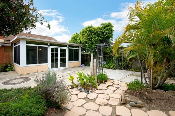 Charming backyard oasis features lush greenery, a stone pathway, and a bright sunroom, perfect for relaxation.