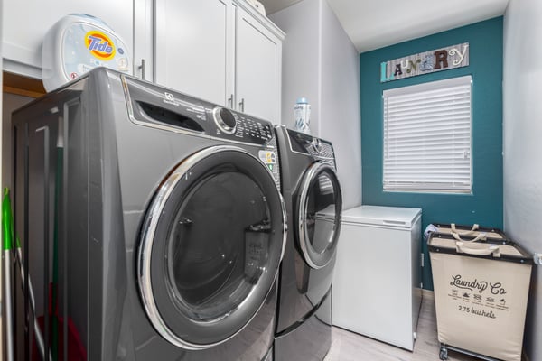 This dedicated laundry room features upper cabinetry for storage, modern laundry appliances, and a window providing natural light. A practical and organized utility space enhances everyday convenience.