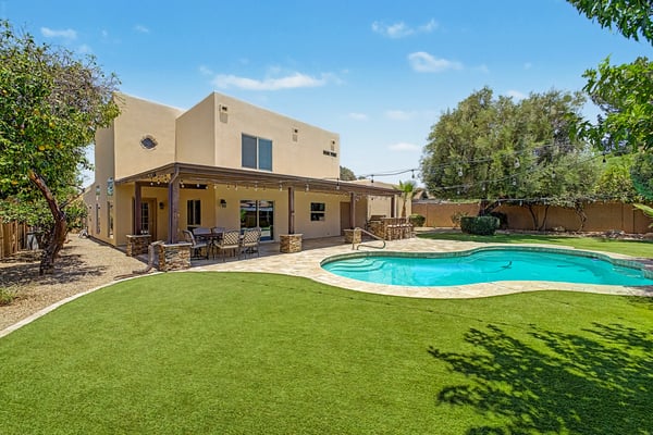Resort-inspired Scottsdale backyard featuring a sparkling swimming pool, expansive artificial turf, mature trees, travertine hardscaping, and a spacious covered patio with outdoor entertaining amenities.