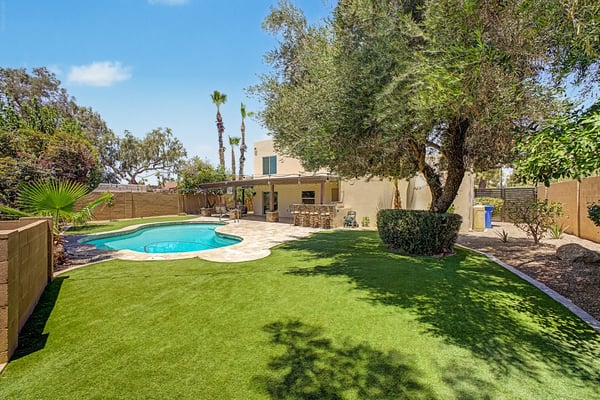 Professionally landscaped Scottsdale backyard with mature shade trees, artificial turf, sparkling pool, covered patio, and outdoor kitchen. An inviting setting for entertaining and relaxing outdoors.