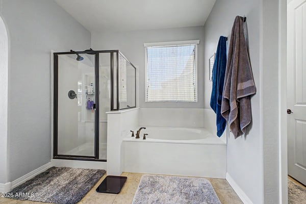 Featuring a soaking tub, separate walk-in shower, tile flooring, and bright natural light, this spacious primary bathroom delivers comfort and convenience in this Goodyear rental home.