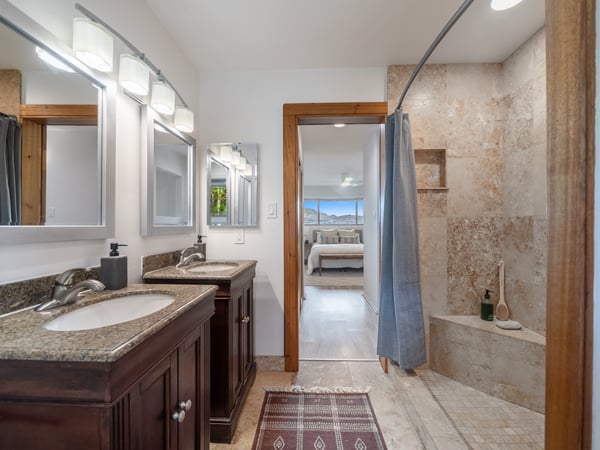 Spacious dual-sink bathroom features a stone-tiled shower and a view of the adjoining bedroom.