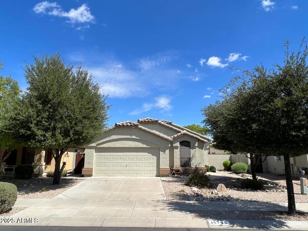 Welcoming front elevation framed by mature trees and desert landscaping for low-maintenance living. The spacious driveway and attached garage provide convenience while complementing the home's appealing design.