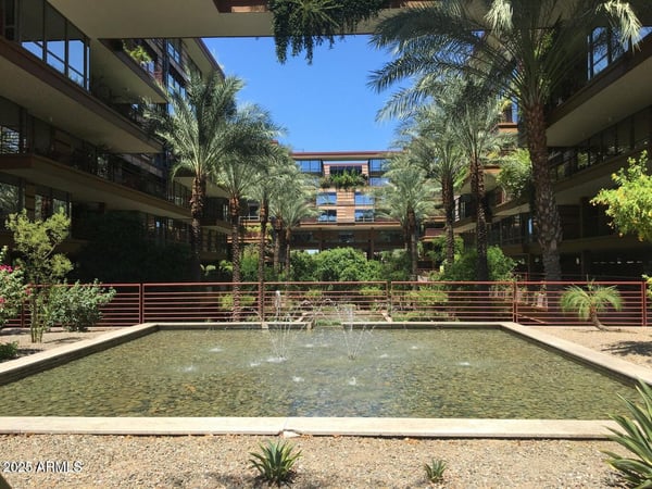 Scenic courtyard showcasing a tranquil fountain, mature palm trees, and lush desert landscaping framed by the iconic architecture of Optima Camelview Village. This peaceful gathering space highlights the community's luxury resort-style atmosphere.