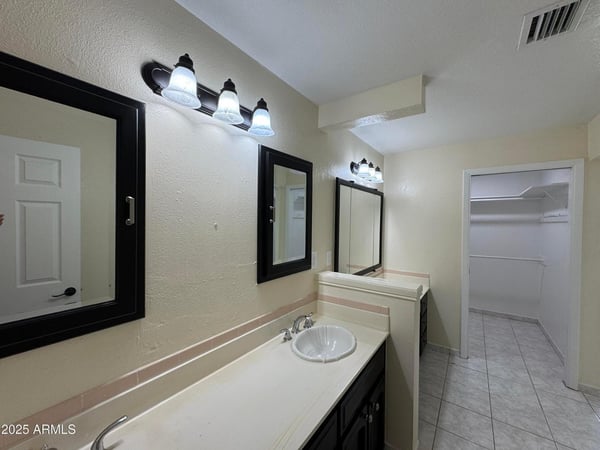 Functional primary bathroom featuring dual sinks, updated lighting, ample storage, and convenient access to a spacious walk-in closet. The thoughtful layout supports both comfort and everyday efficiency.