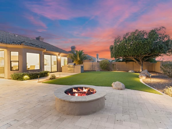Twilight backyard featuring a glowing fire pit, outdoor kitchen, professionally landscaped yard, and spacious travertine patio. Experience luxury outdoor living in Scottsdale.