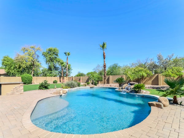 Wide view of the sparkling swimming pool showcasing the waterfall feature, baja shelf, paver decking, mature landscaping, and generous entertaining areas in this Glendale property.